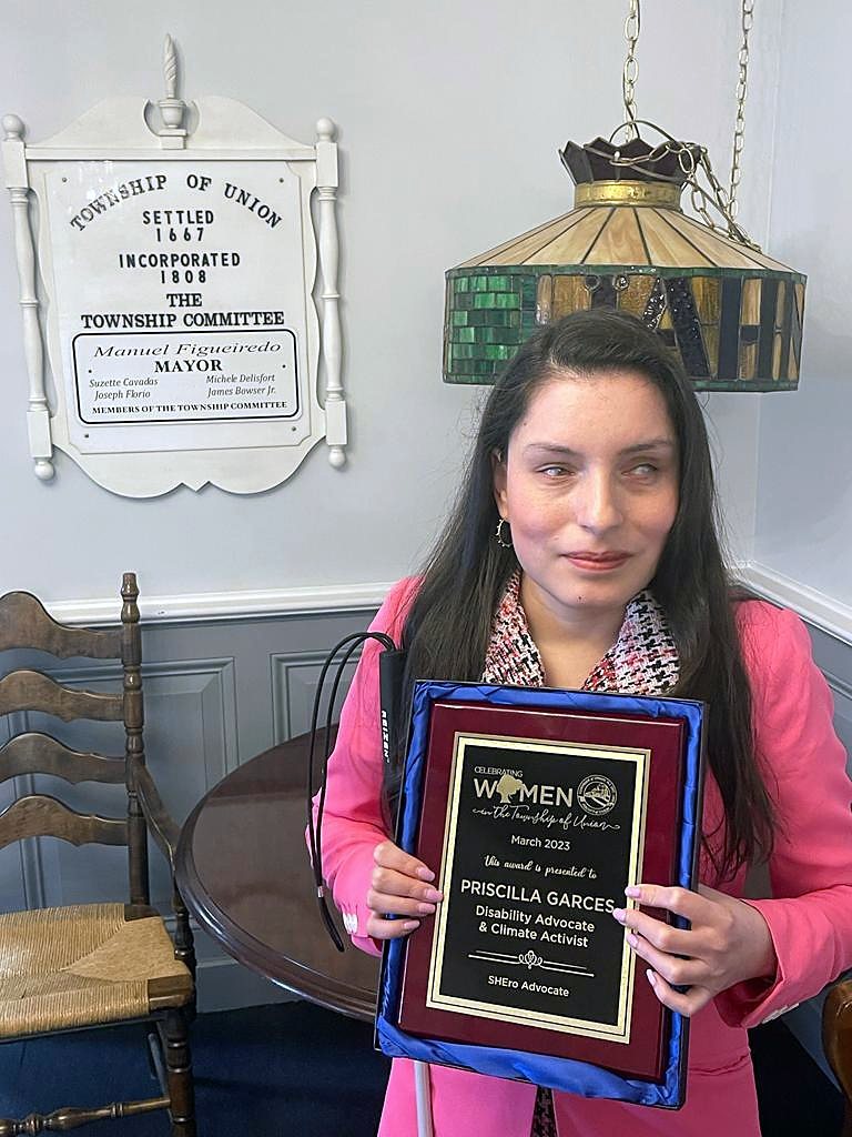 Priscilla Garces in a pink blazer holds a framed award from the Township of Union, Celebrating Women, March 2023, naming her Disability Advocate and Climate Activist and SHEro Advocate. Her white cane rests at her side.