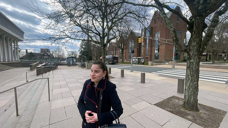 Priscilla Garces in a navy coat, smiling, standing on a stone plaza with bare trees and brick buildings behind her.