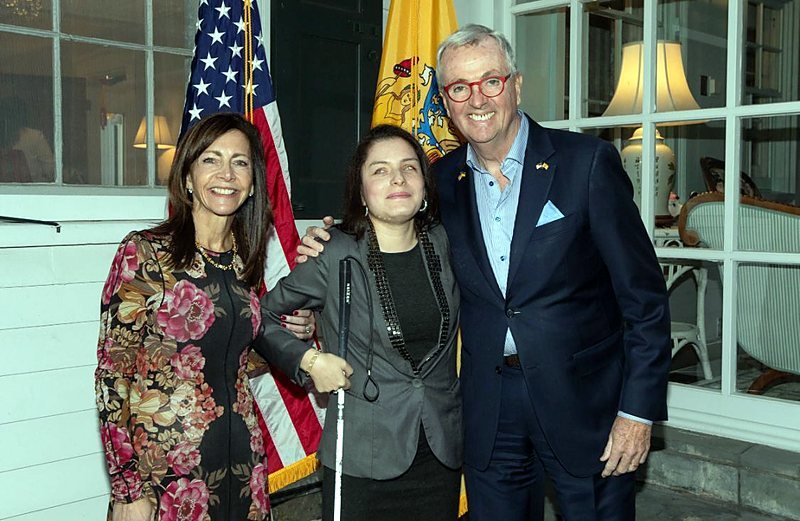 Priscilla Garces, holding a white cane, stands between New Jersey Governor Philip D. Murphy and another guest, in front of the American and New Jersey flags.
