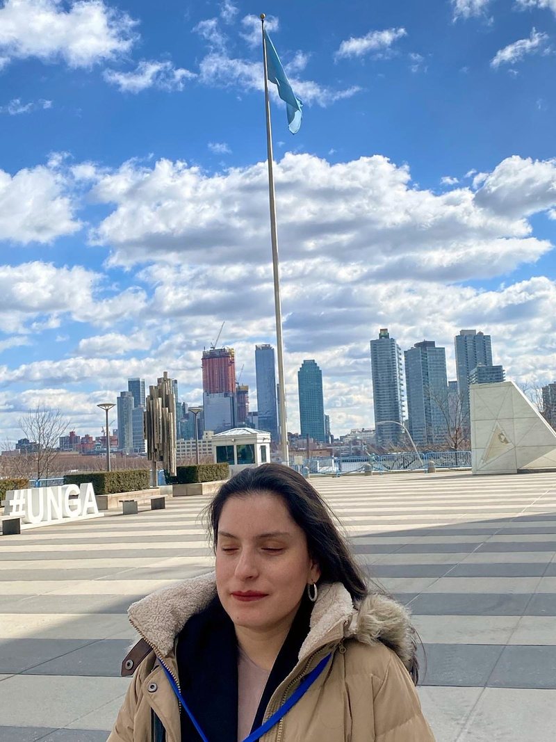 Priscilla Garces in a winter coat with a blue lanyard, standing on the plaza at United Nations Headquarters, with the UN flag, the #UNGA sign and the Manhattan skyline behind her.